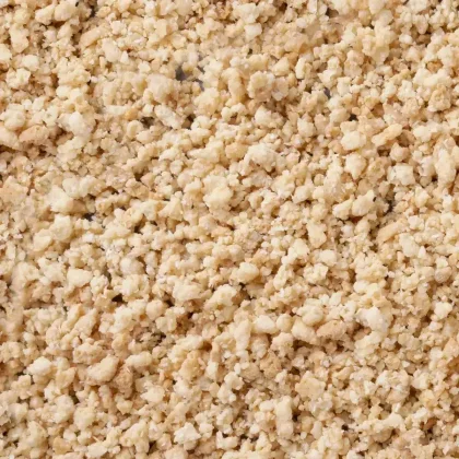 Detailní záběr na světle béžové, drolivé granule s hrubou, drobivou texturou pokrývající celý záběr.